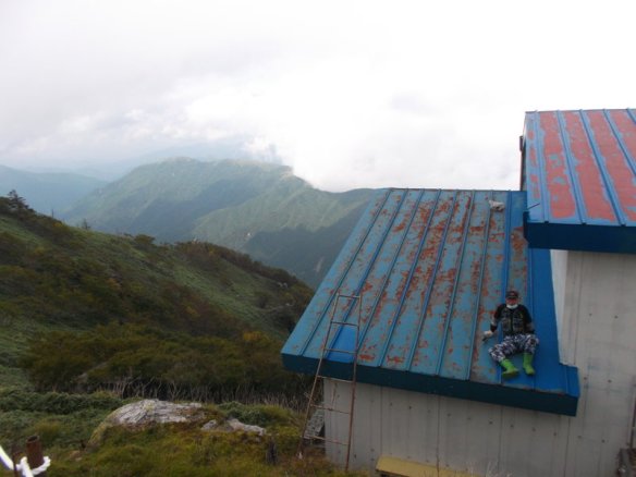 In the background, there is a blanket of white cloud smothering green mountains.  In the foreground, on the right side, is the blue metal roof of a building, and there is a man sitting on the roof.  The man looks small compared to the roof, let alone the mountains.